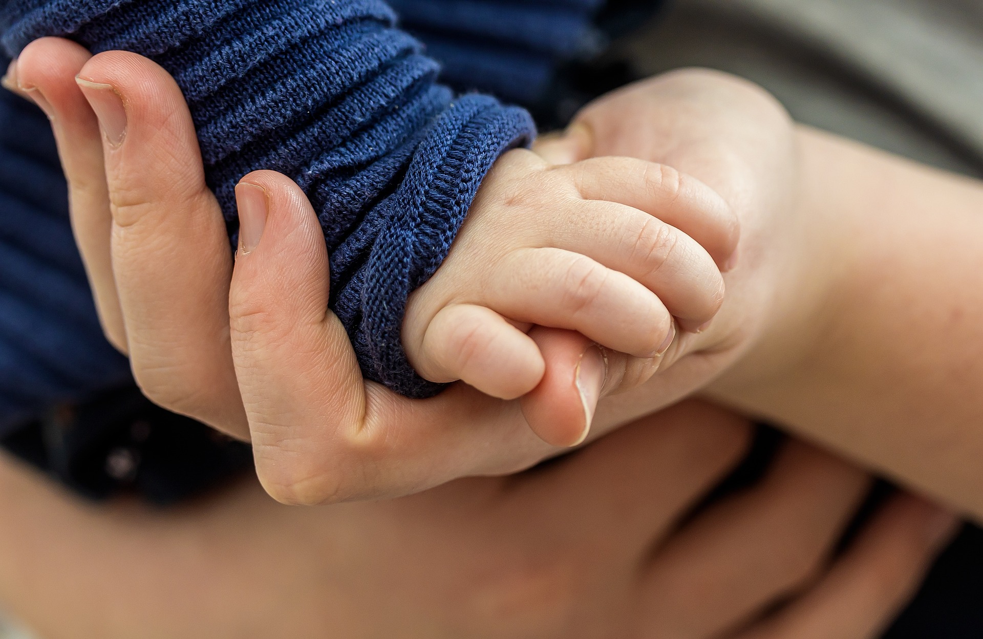 A baby's hand grasps that of their parents, close up. They both have light skin and the baby wears a long sleeved jumper.